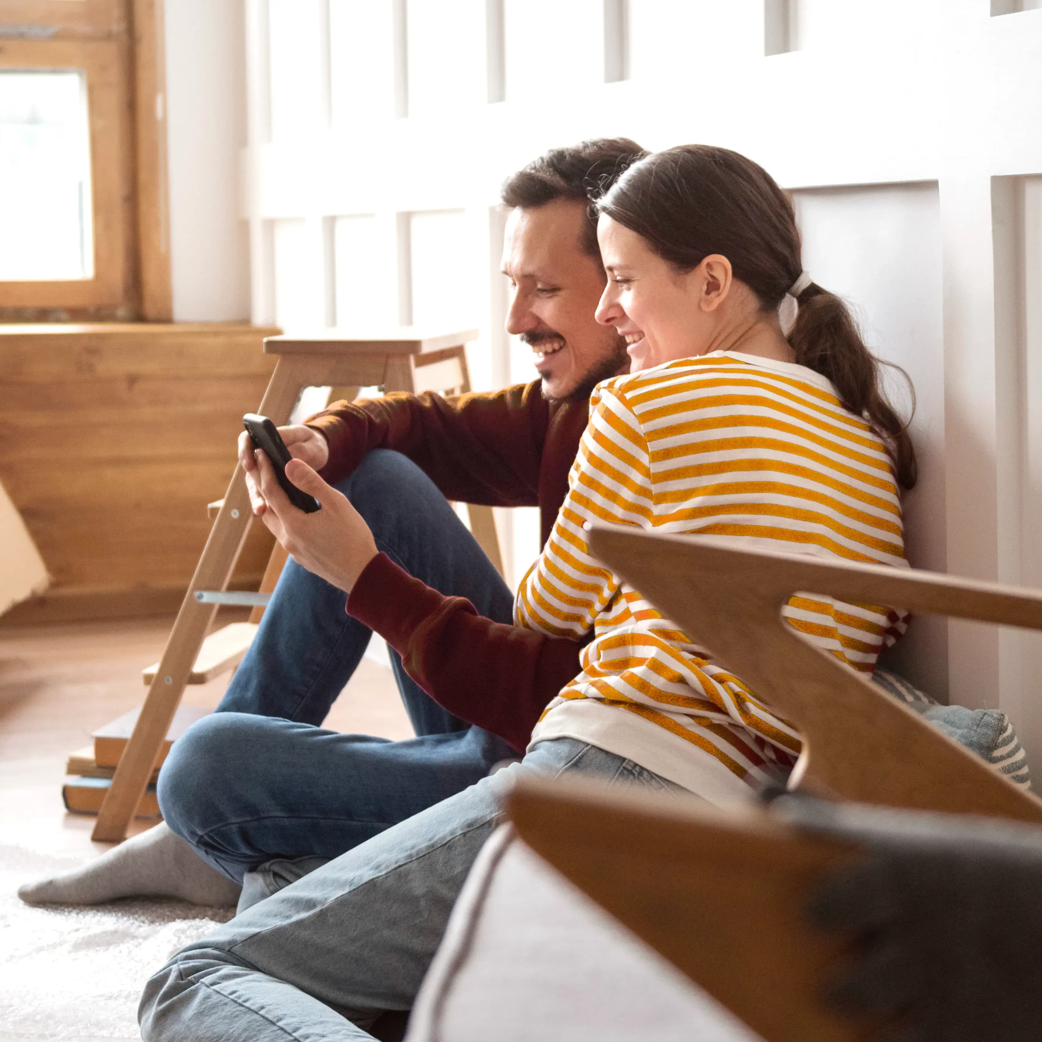 Kobieta i mężczyzna siedzą na podłodze w nowo urządzanym mieszkaniu, razem patrzą na ekran smartfona.