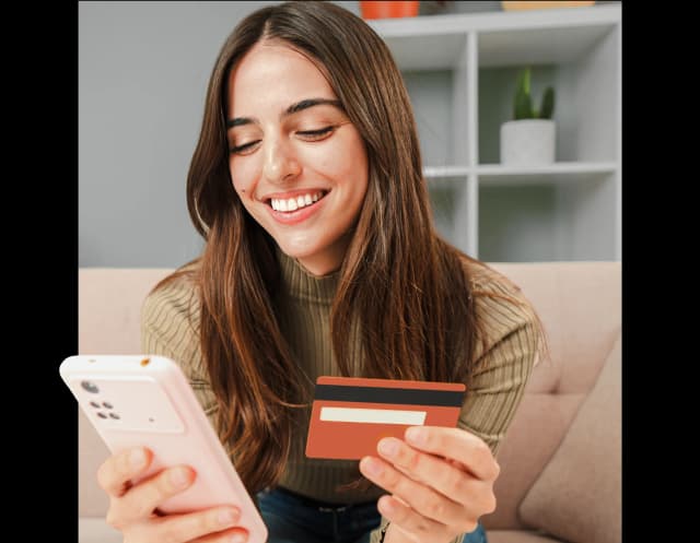 Uśmiechnięta kobieta siedzi na kanapie, trzyma kartę kredytową i patrzy na ekran telefonu.