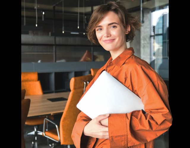 Uśmiechnięta kobieta w pomarańczowej koszuli stoi w biurze, trzyma laptopa.