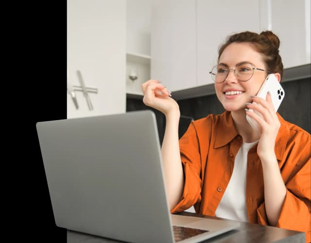 Uśmiechnięta kobieta w okularach rozmawia przez telefon, siedząc przy laptopie.