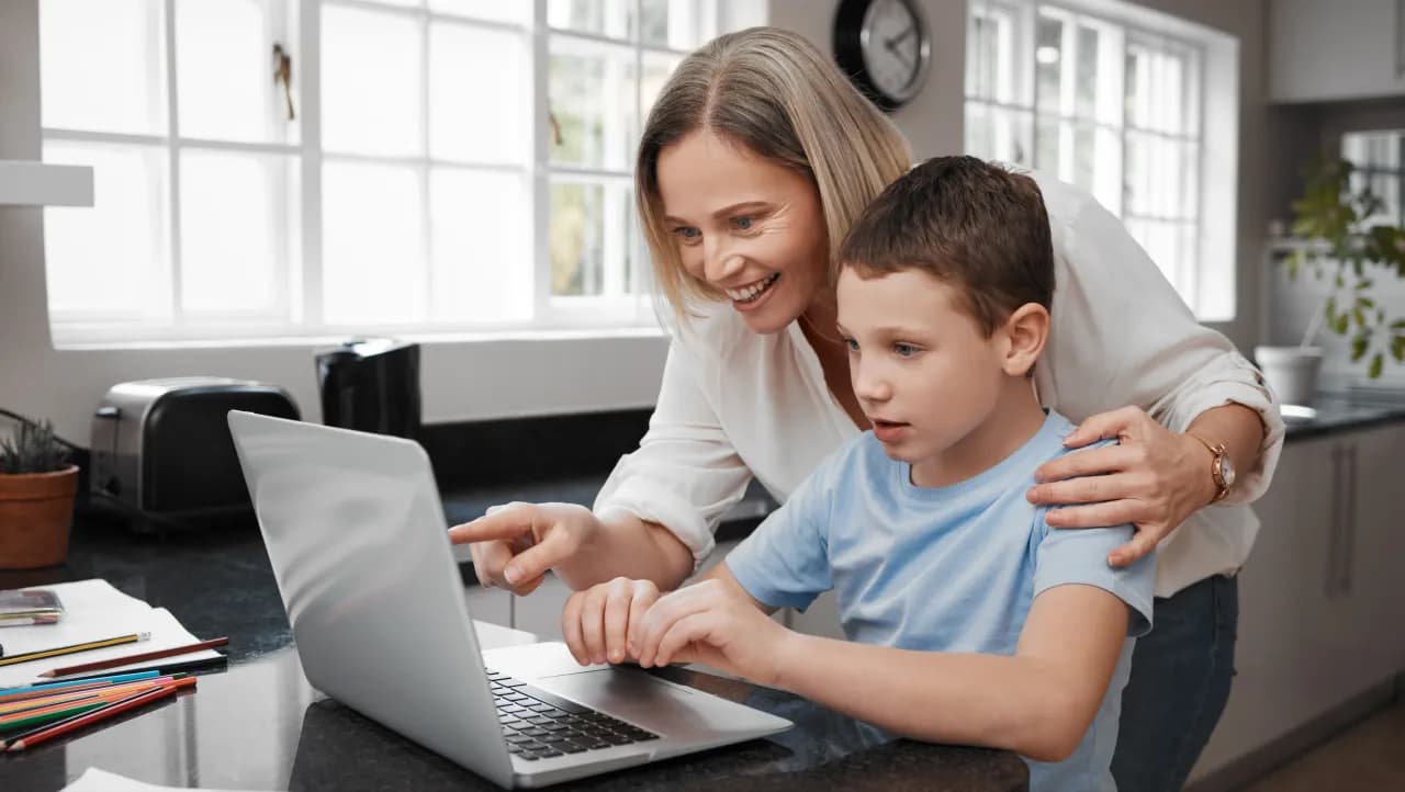 Mama stoi obok syna, który siedzi na krześle przy stole. Obydwoje patrzą w ekran laptopa. Mama wyszukuje na stronie informacje.