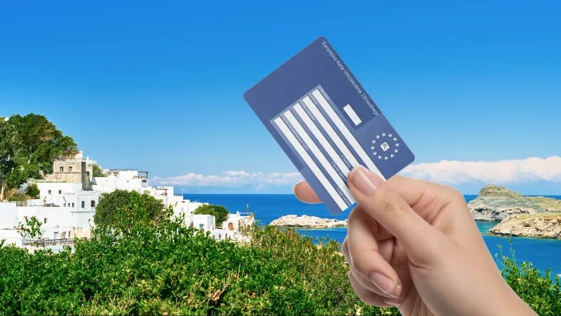 Dłoń trzyma Europejską Kartę Ubezpieczenia Zdrowotnego (EKUZ) na tle nadmorskiego krajobrazu z białą zabudową, zielenią i turkusowym morzem pod bezchmurnym niebem.