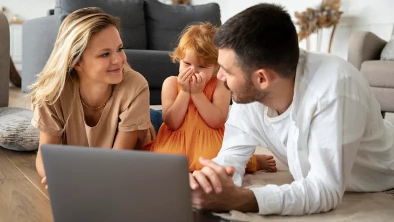 Młoda kobieta, dziecko i mężczyzna leżą na podłodze w domu. Uśmiechają się. Przed nimi stoi otwarty laptop. 