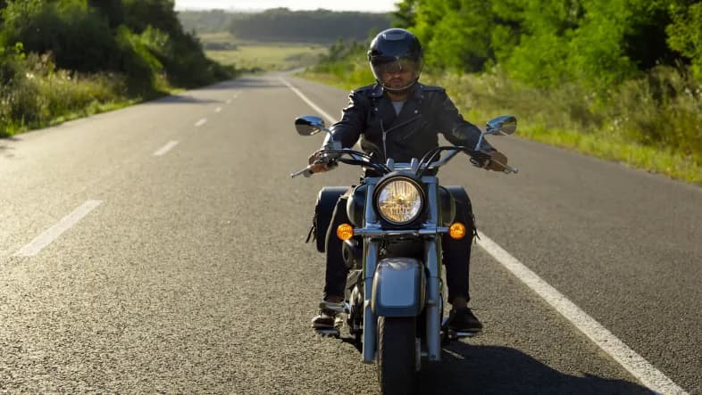 Mężczyzna jedzie drogą dwupasmową na motocyklu typu chopper. Ma na sobie kask i czarną skórzaną kurtkę.