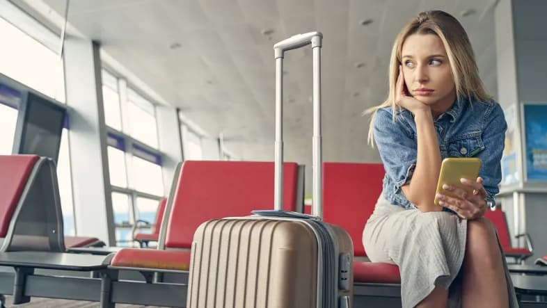 Kobieta siedzi smutna na fotelu na lotnisku. W ręku trzyma telefon. Obok niej stoi walizka. Kobieta ubrana jest w koszulę koloru niebieskiego oraz beżową spódnicę. Prawą ręką podpiera głowę, patrzy w kierunku szyb, za którymi są pasy startowe.