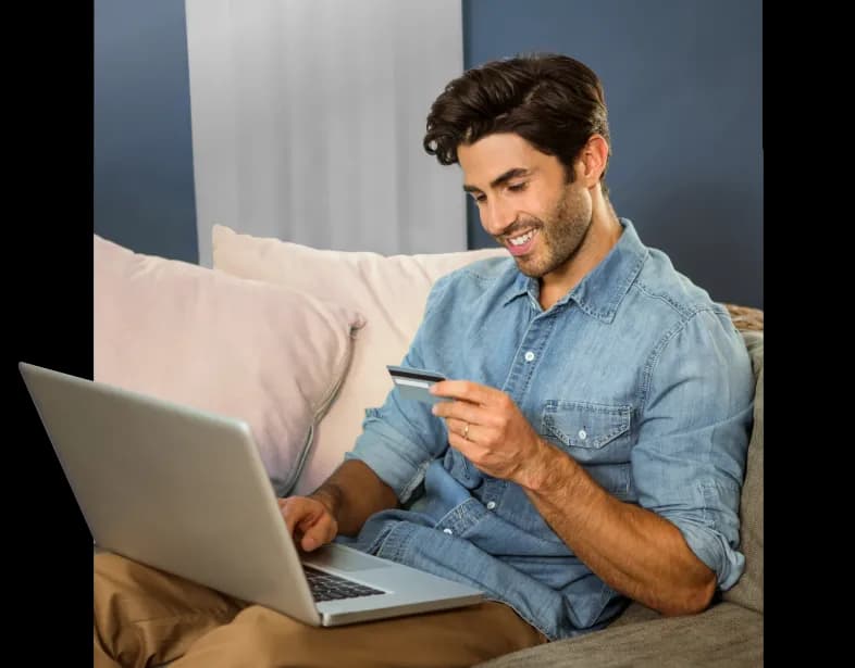 Uśmiechnięty meżczyzna trzyma kartę kredytową i korzysta z laptopa.