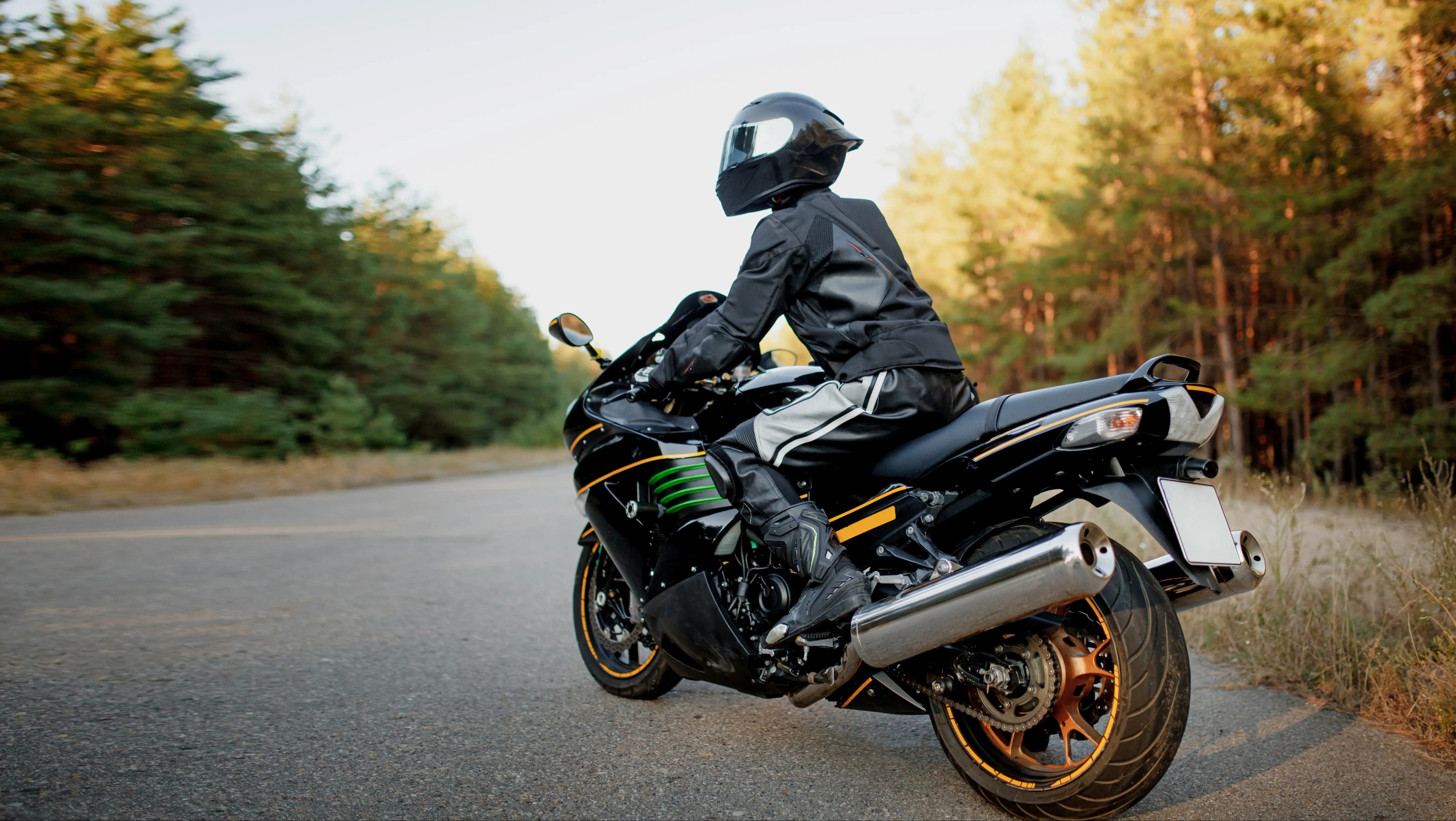 Mężczyzna siedzi na sportowym motocyklu znajdującym się na drodze. Ubrany jest w czarną kurtkę, spodnie, buty motocyklowe, na głowie ma czarny kask. Znajduje się na asfaltowej drodze przebiegającej przez las.