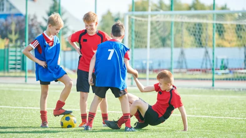 Czterech chłopców w strojach sportowych stoi na boisku szkolnym. Jeden z chłopców kopie piłkę.