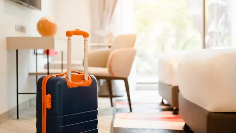 na pierwszym planie widoczna jest walizka stojąca w pokoju hotelowym, na drugim planie widać pokój hotelowy - łóżko, szafki oraz okno